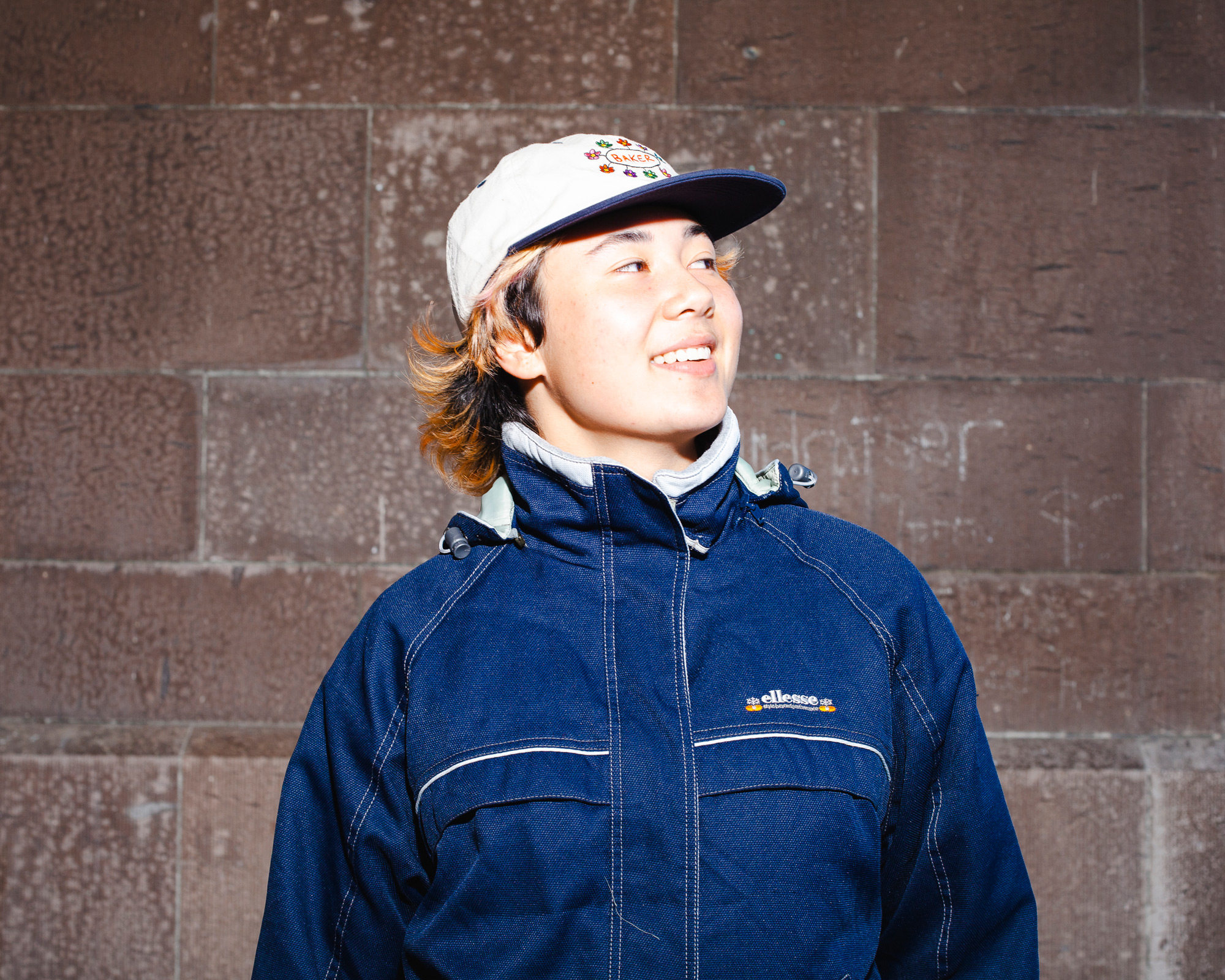 Portrait photograph of Gala Baumfield standing in front of a brick wall. She is looking away off camera and smiling.