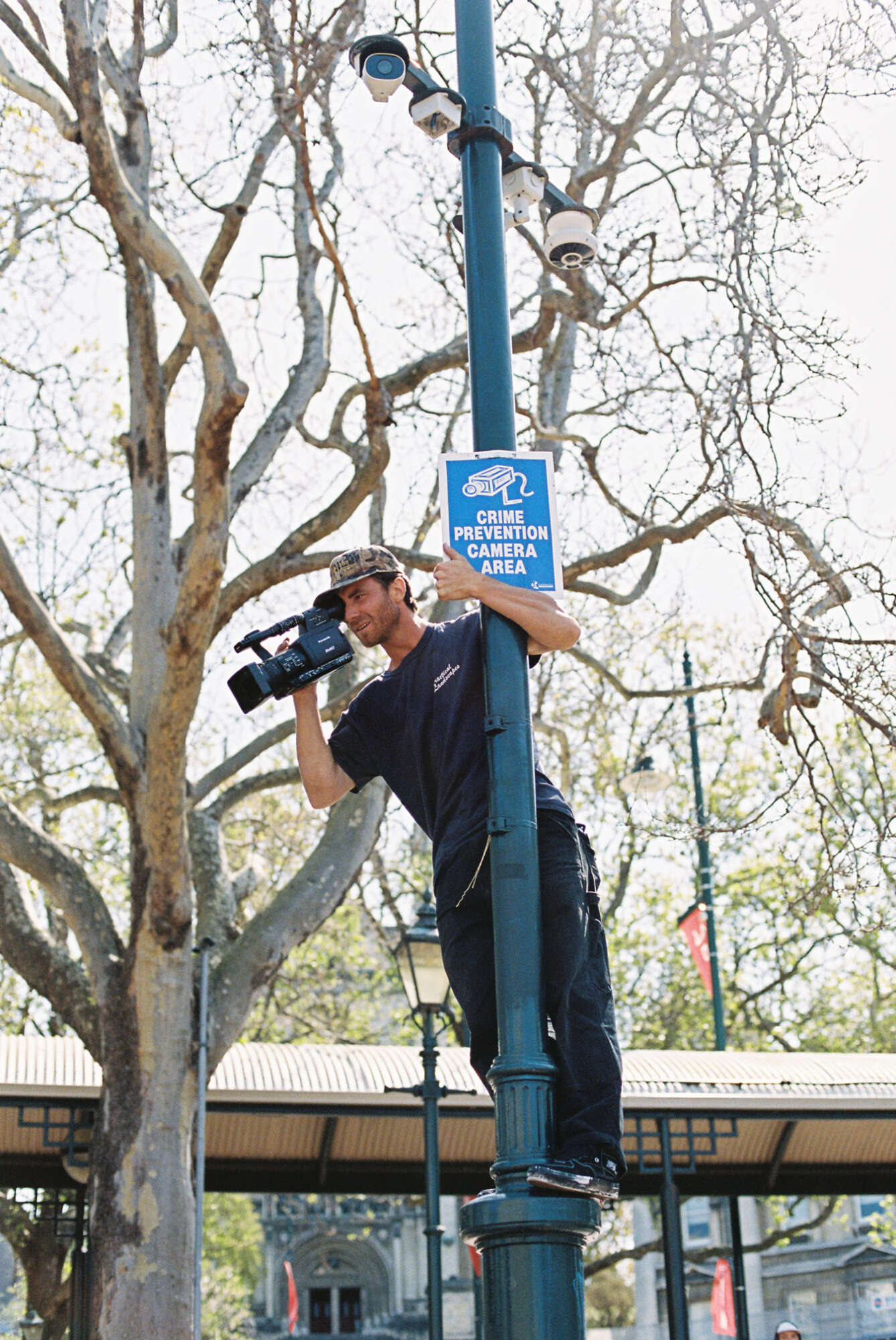 A filmer hanging from a lamp post with a sign reading security camera in action.