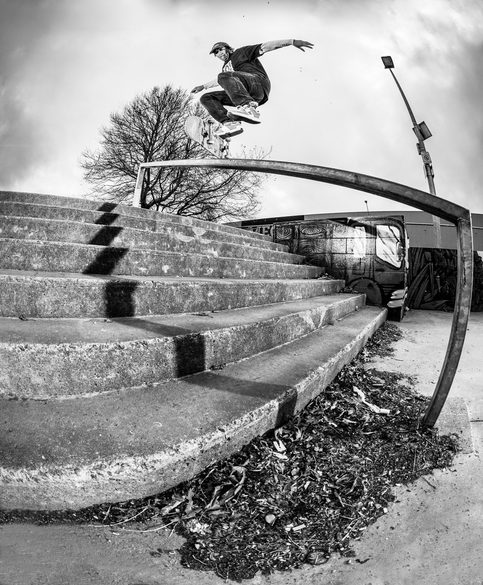 Black and white photo of Pauly Kauri doing a kickflip frontside boardslide at Ashburton Skatepark. The image is shot using a fisheye lens and is pointing up at the rail and Pauly from the ground.