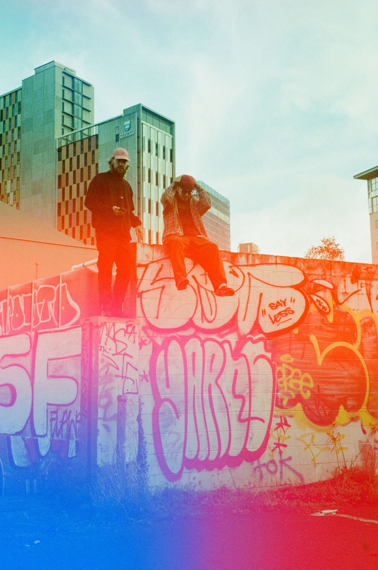 Members of the hip-hop group No Comply sitting on a wall in an urban environment. There's graffiti on the wall. The photo has a light leak and grainy quality to it.