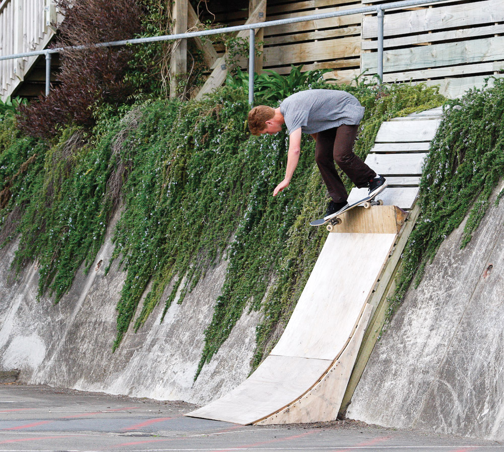 Tim Watson riding a skateboard and doing a backside Smith grind on a DIY quarterpipe position on a banked retaining wall at Wellington Hospital in Newtown.